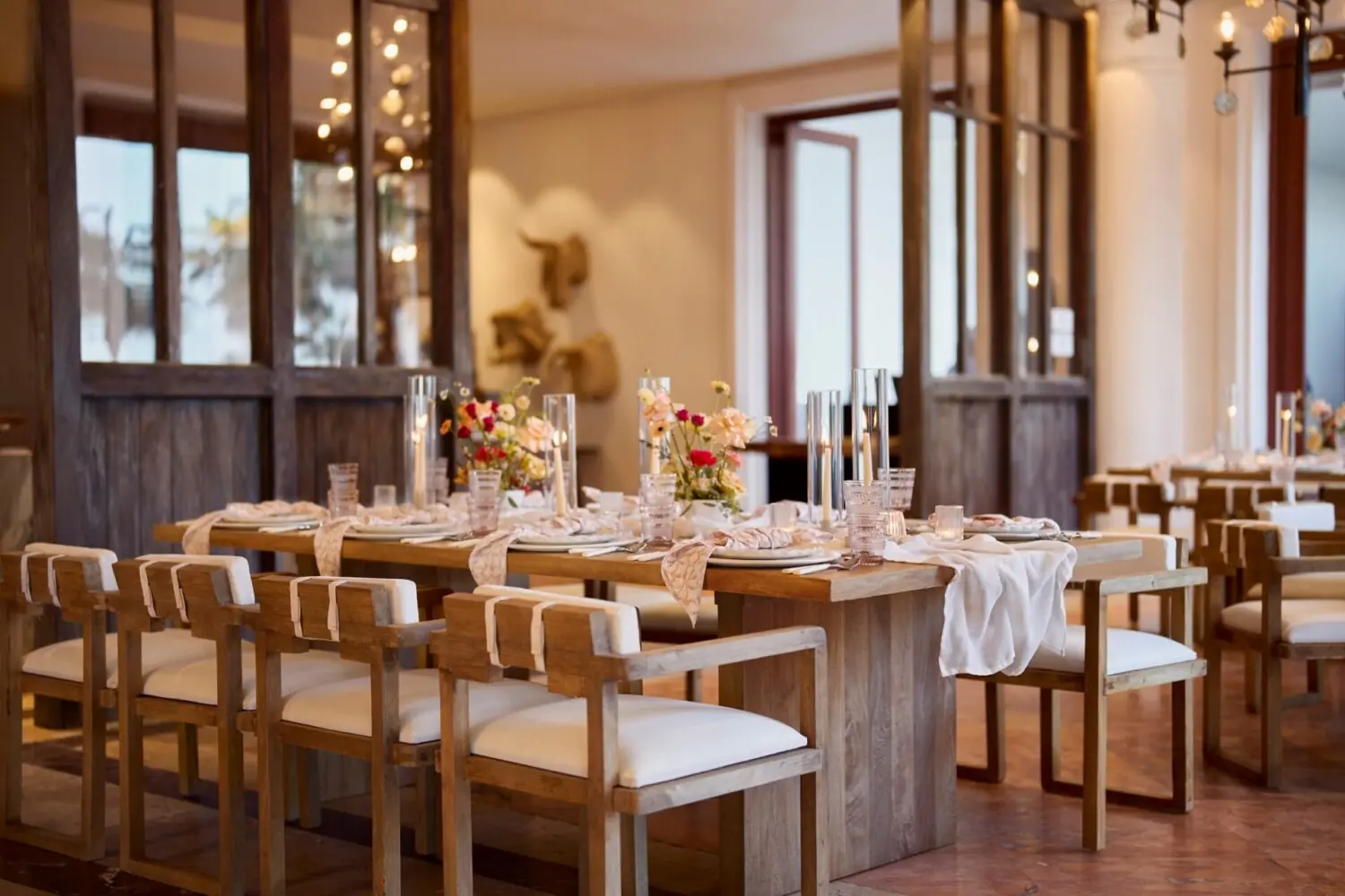 Rustic wood dining tables with white chairs, elegant table settings, florals, candles, warm lighting, indoor ambiance.