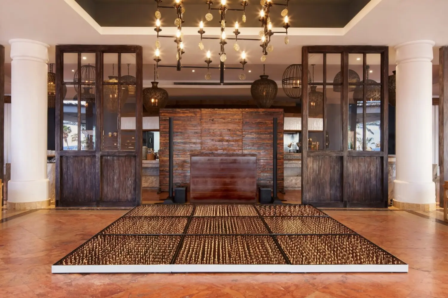 Indoor venue with a dark wooden DJ booth, sparkling light-up dance floor, chandeliers, columns, and rustic decor.