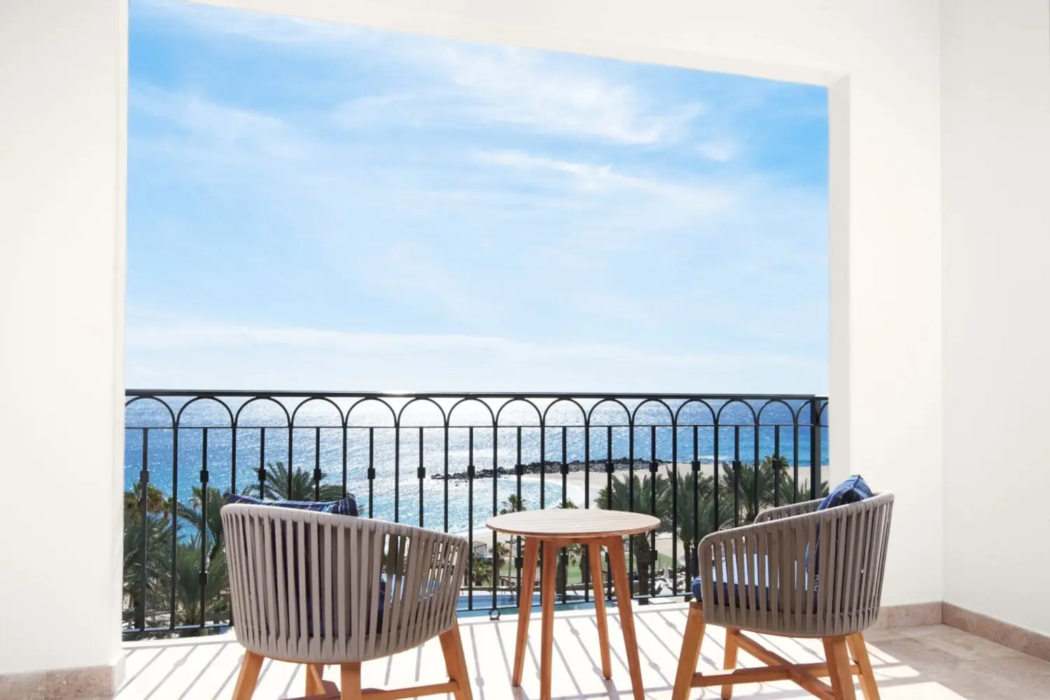Dos sillas y mesa en balcón con vista al mar, palmeras y cielo azul claro.