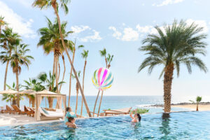 Family plays with colorful beach ball in infinity pool, surrounded by palm trees and ocean.
