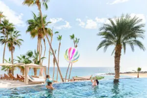 Familia juega con una pelota de playa de colores en una piscina infinita, rodeada de palmeras y océano.