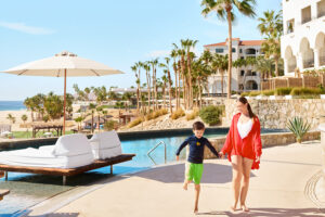 Mother and child walk hand-in-hand by resort poolside, palm trees and ocean in background.