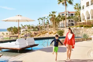 Madre y su hijo caminan de la mano junto a la piscina del resort, con palmeras y el océano de fondo.