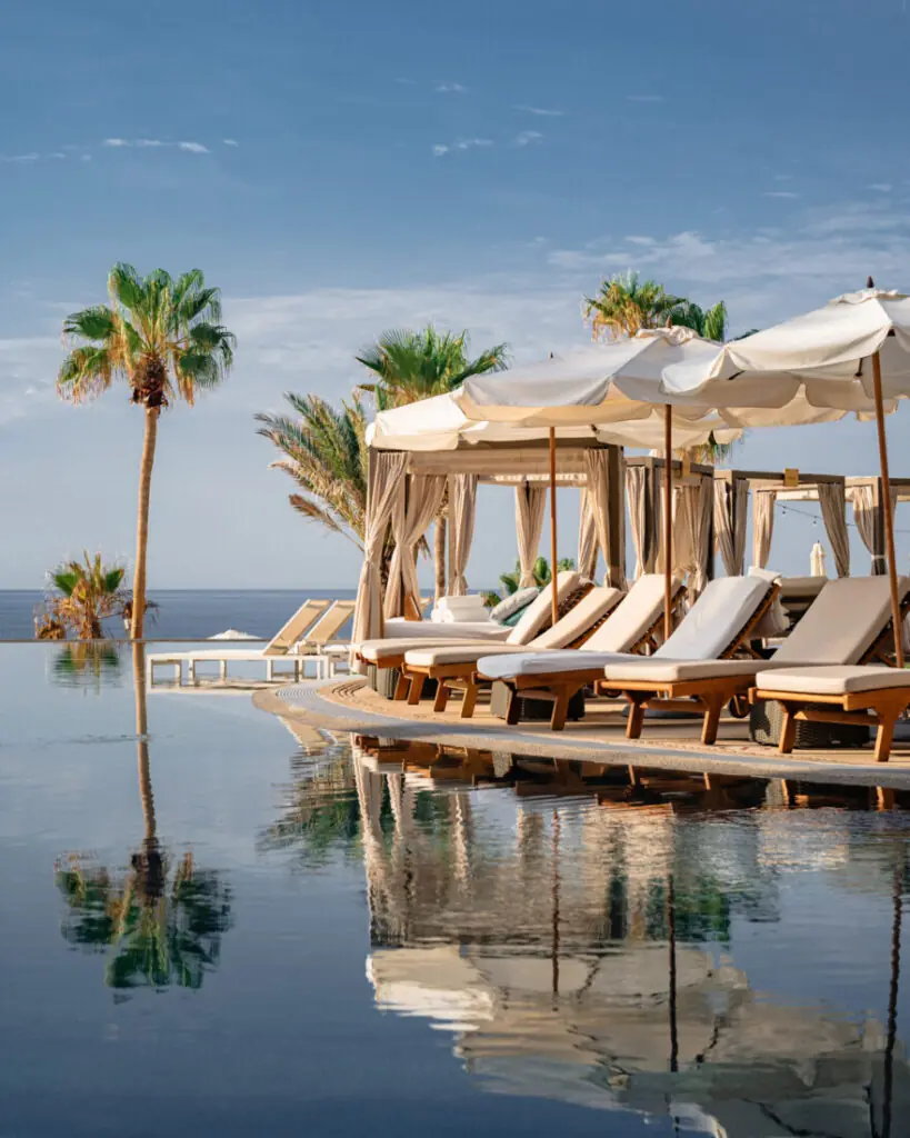 Luxurious poolside cabanas and lounge chairs overlook the ocean, surrounded by palm trees and blue sky.