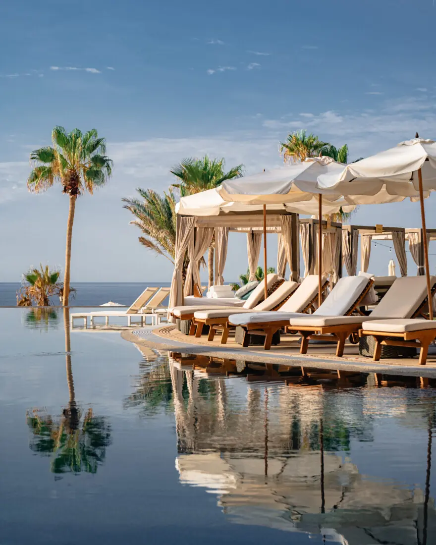 Luxurious poolside cabanas and lounge chairs overlook the ocean, surrounded by palm trees and blue sky.