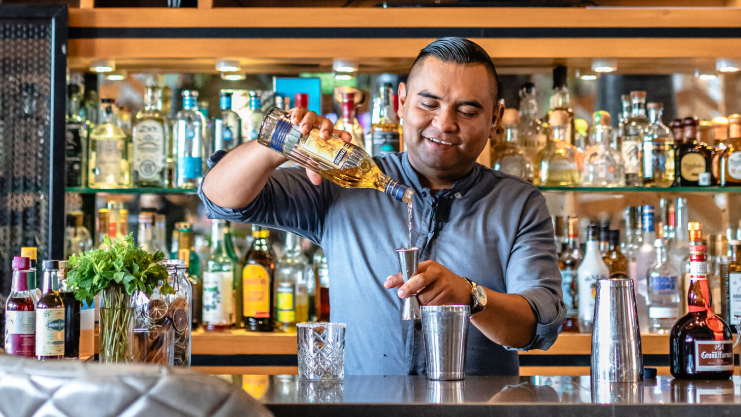 Un camarero sonriente vierte licor en un vaso medidor mientras prepara un cóctel, rodeado de botellas e ingredientes frescos.
