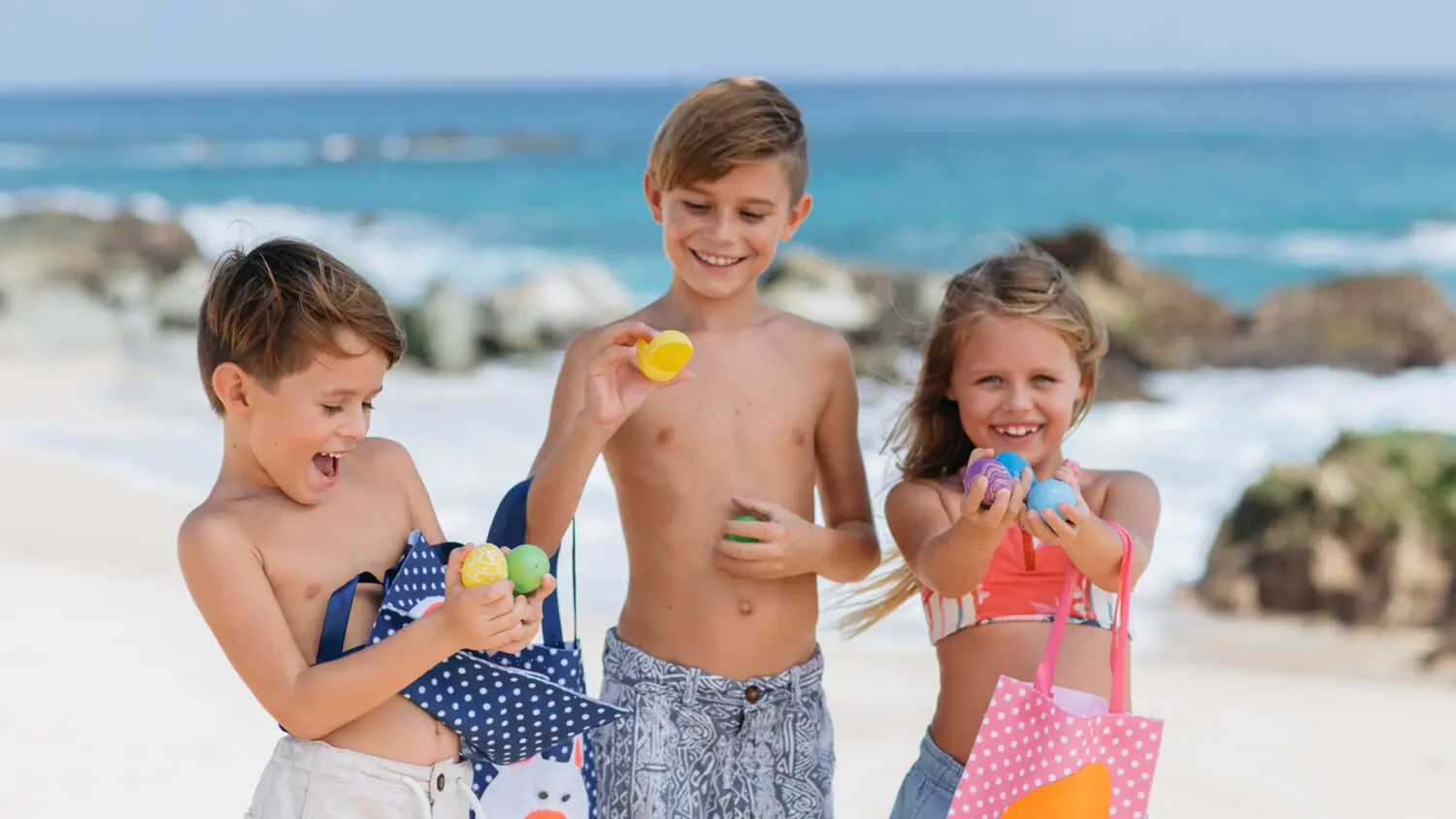 Tres niños sonrientes en la playa sosteniendo coloridos huevos de Pascua, con bolsas de mano y fondo de océano.