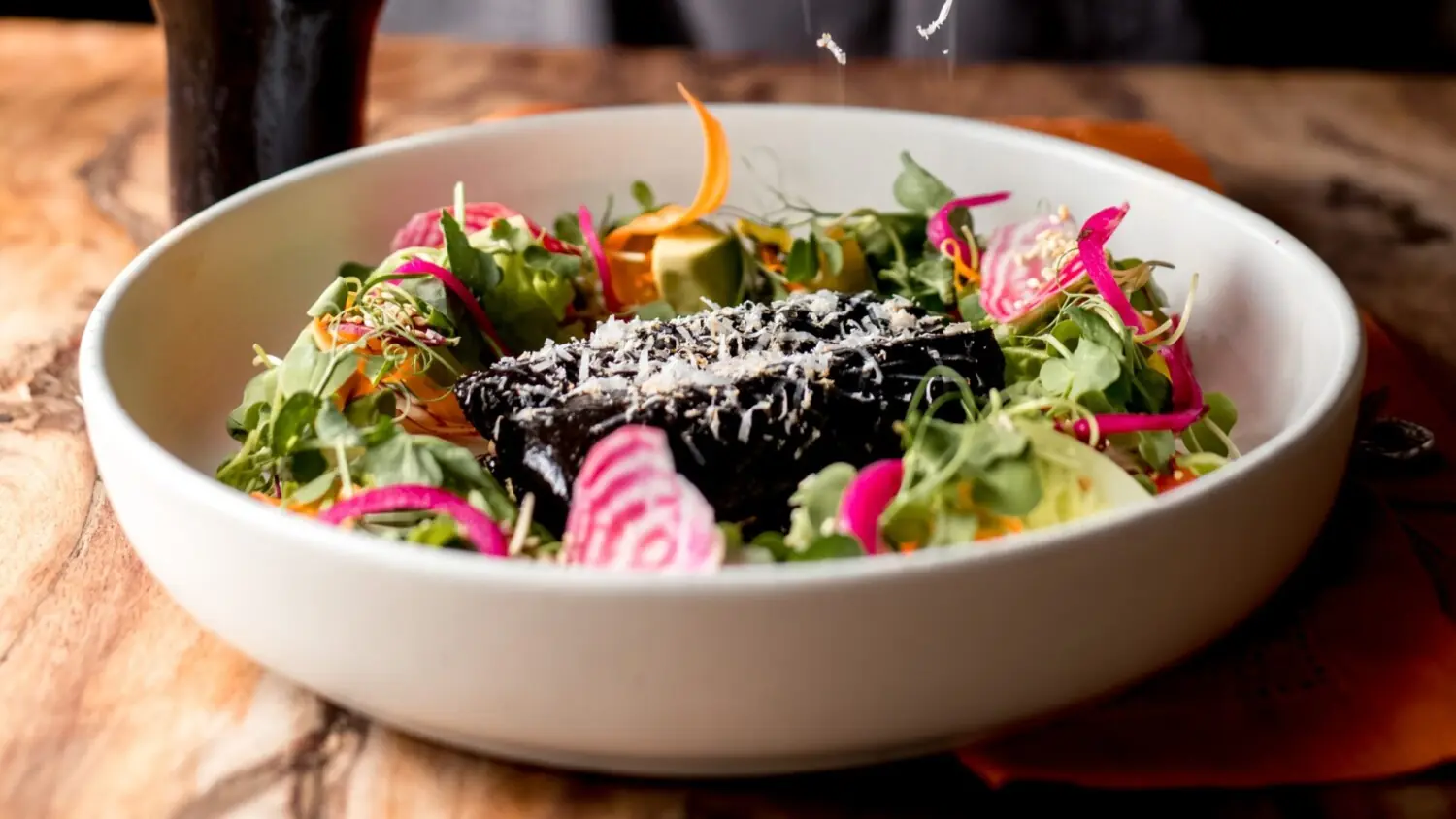 Gourmet dish featuring dark mole centerpiece, surrounded by colorful vegetables, microgreens, and shredded cheese garnish.