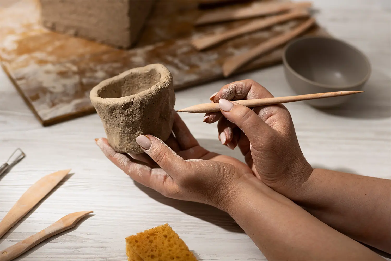 Manos dando forma a una pequeña olla de barro con una herramienta de escultura de madera, rodeadas de instrumentos de cerámica y esponja.