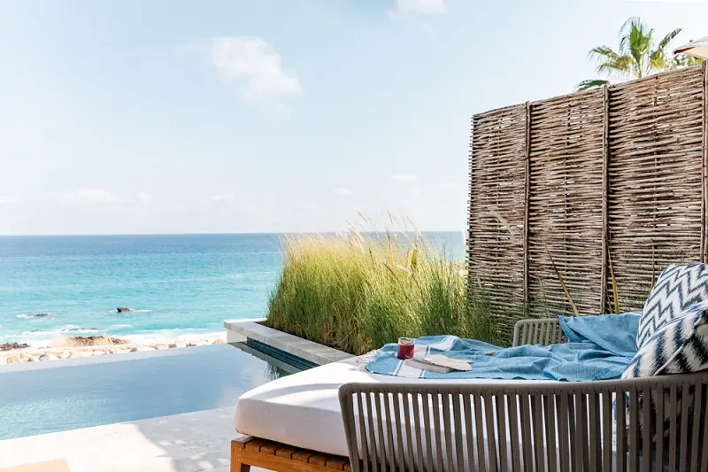 Cama tipo lounge frente al mar junto a una piscina infinita privada, con mampara de privacidad tejida, vegetación y vista al mar azul claro.