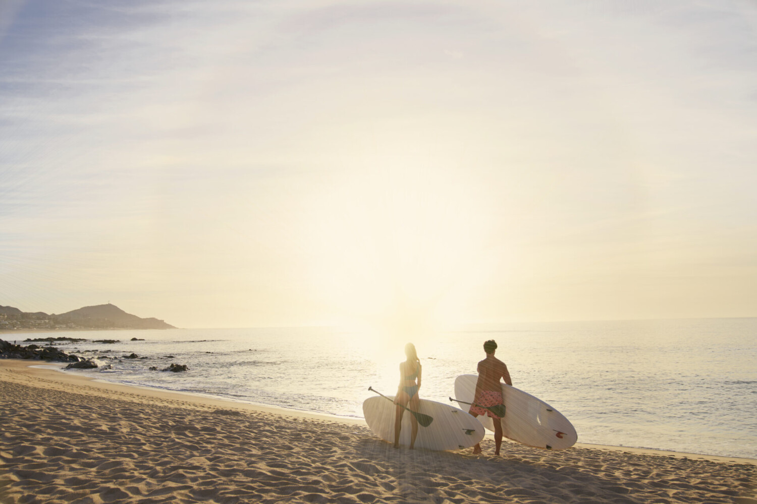 Dos personas llevando tablas de surf en la playa de arena al amanecer, frente a las olas del océano con un horizonte dorado.
