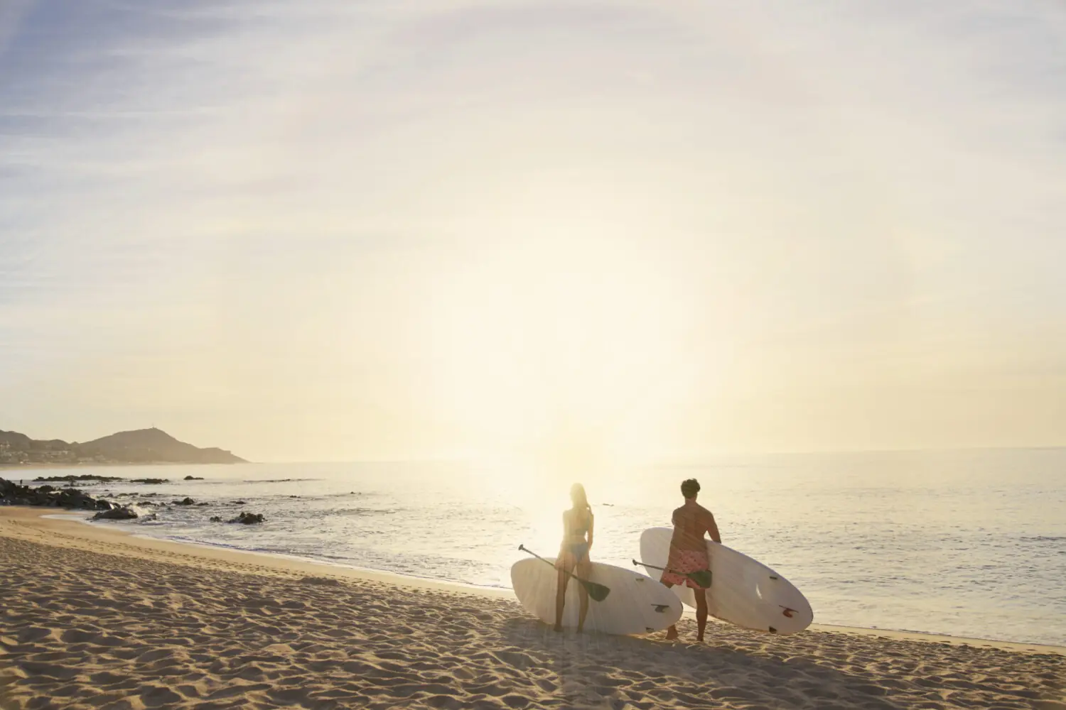 Dos personas llevando tablas de surf en la playa de arena al amanecer, frente a las olas del océano con un horizonte dorado.