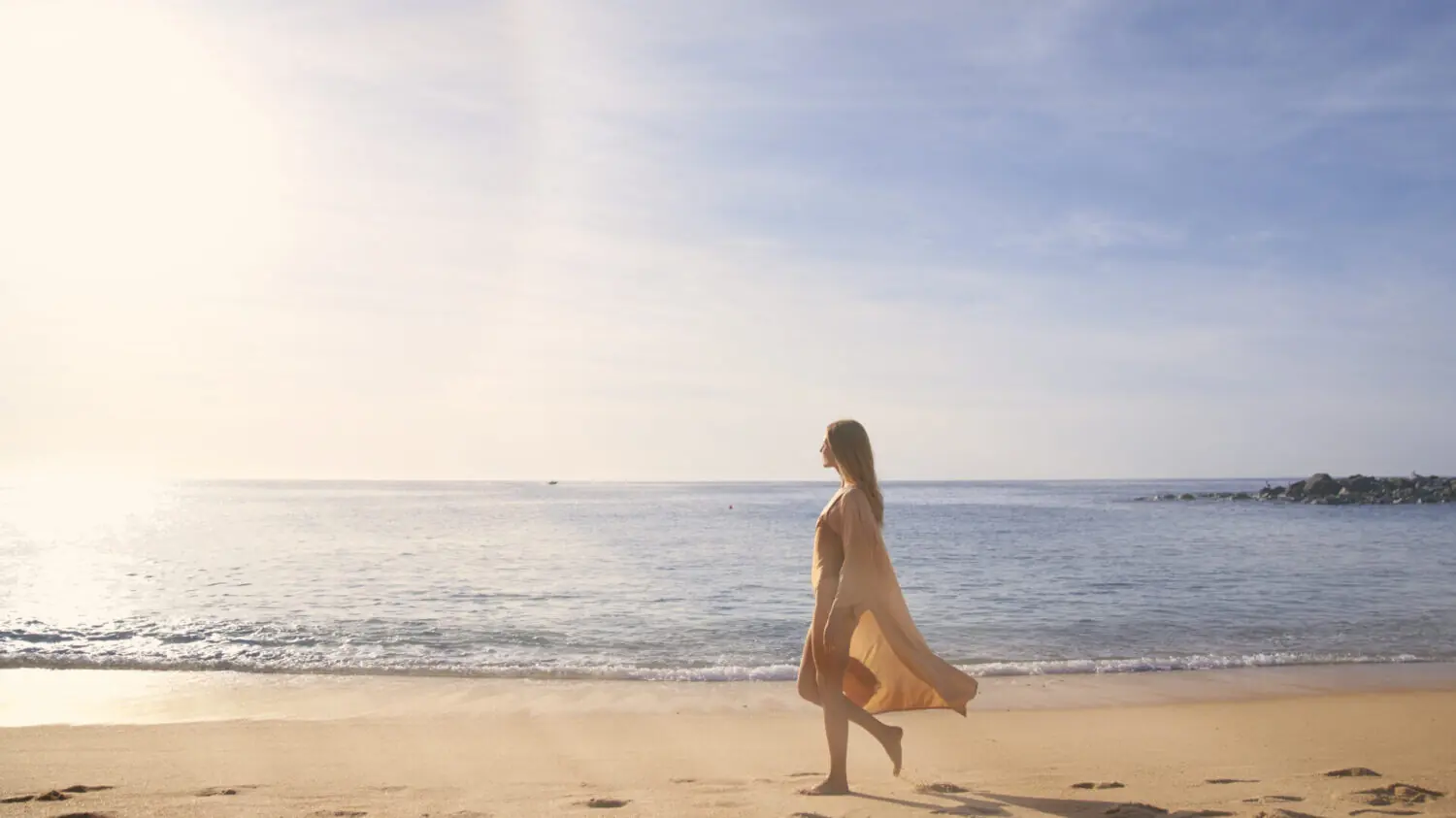 Woman in flowing dress walking on sandy beach beside calm ocean under clear blue sky at sunrise.