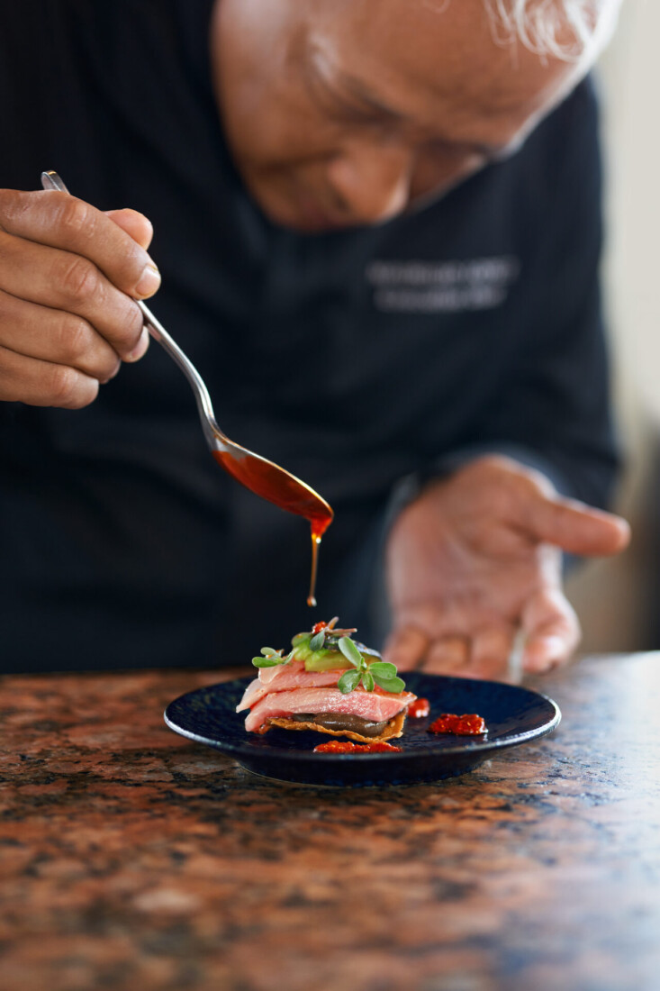 Chef carefully plating a gourmet dish, drizzling sauce over fresh tuna with microgreens on a dark plate.
