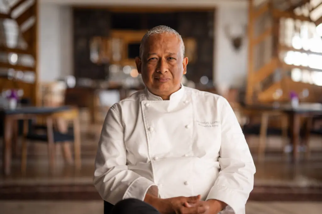 Chef in a white uniform seated in a restaurant dining room, looking confidently at the camera.
