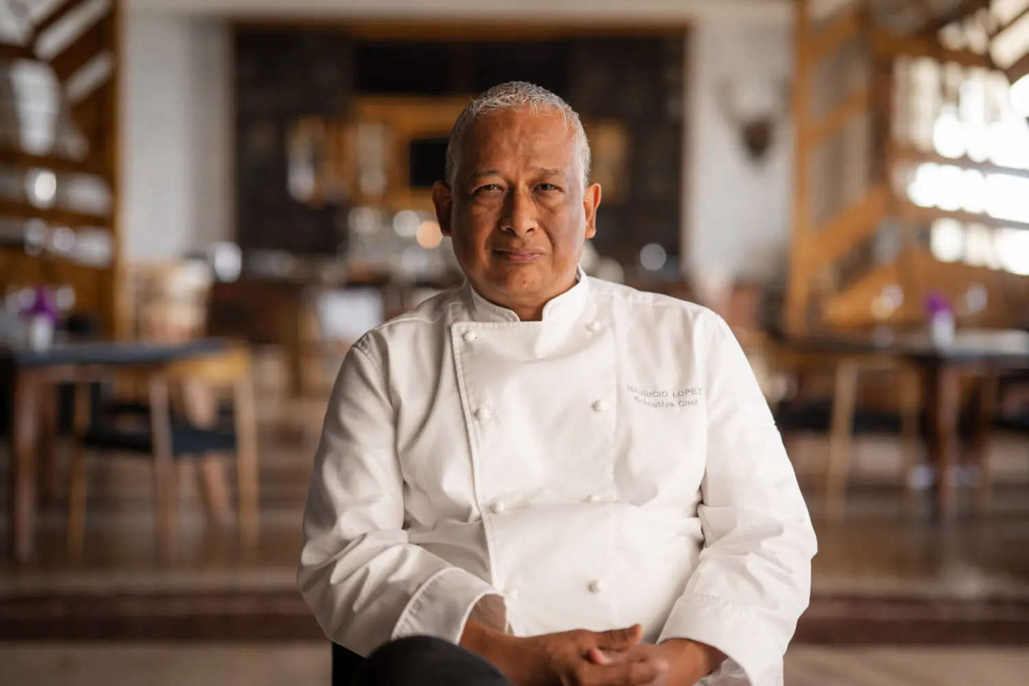 Chef in a white uniform seated in a restaurant dining room, looking confidently at the camera.