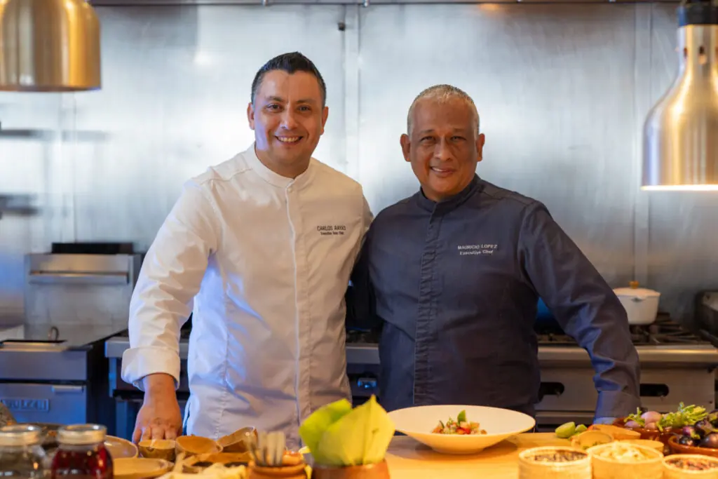 Two chefs smiling in a professional kitchen, standing behind a counter with plated food and ingredients.