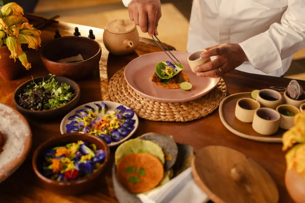 Chef carefully plating a gourmet dish with edible flowers and sauce on a wooden table.
