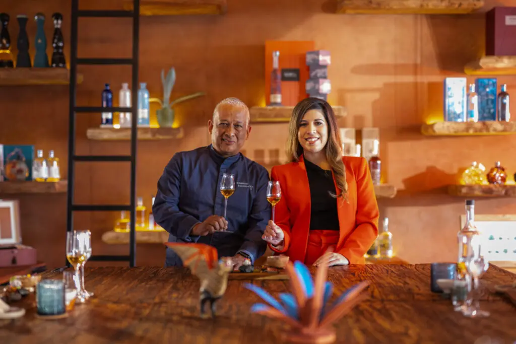 Two people holding tasting glasses at a stylish bar with shelves of bottles and warm ambient lighting.