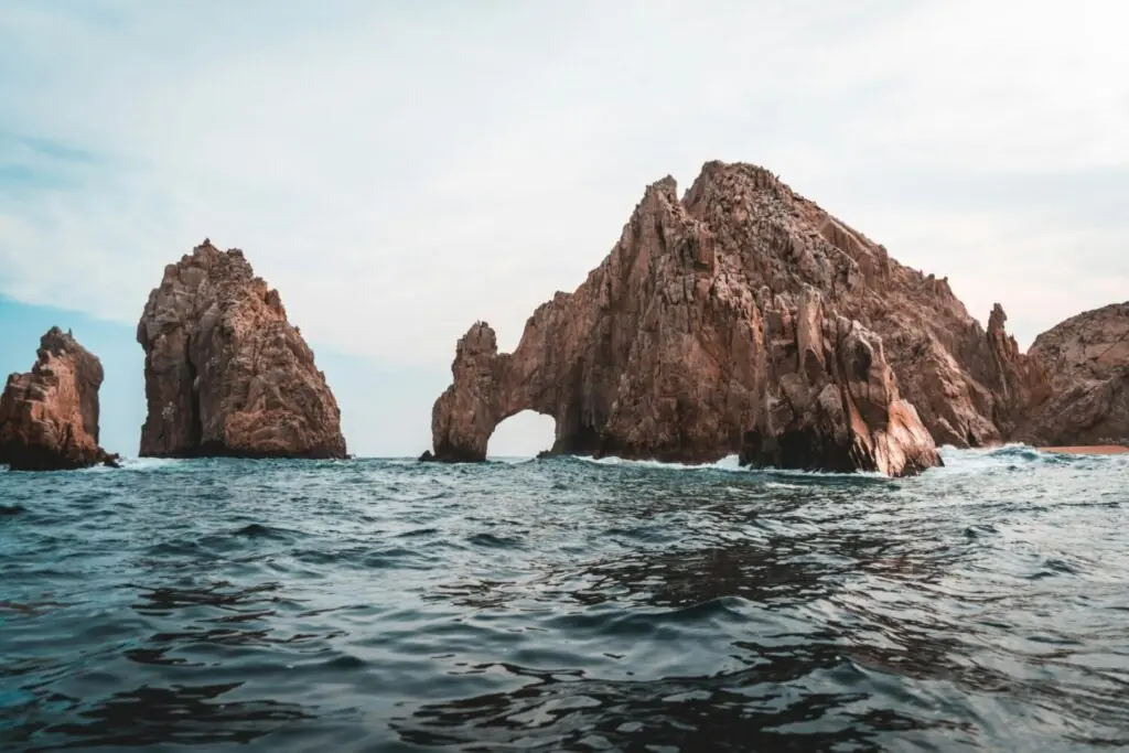 Rocky sea arch and jagged cliffs rising from the ocean under a pale sky, viewed from the water.