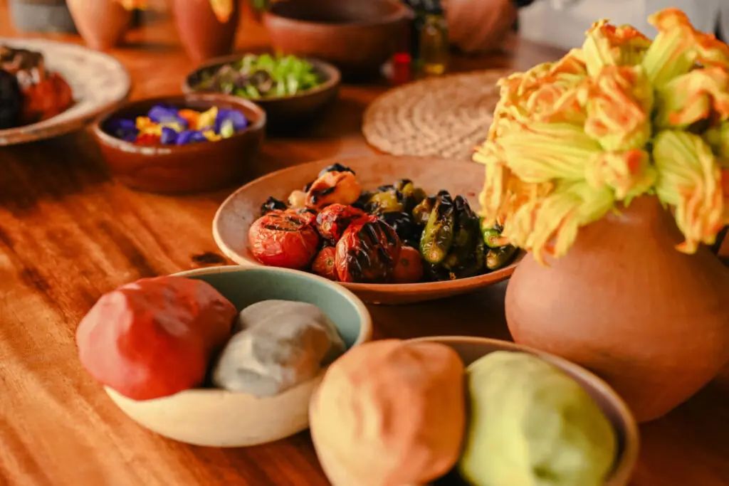 Colorful rustic table spread with clay bowls, fresh dough, roasted vegetables, and squash blossoms in warm light.
