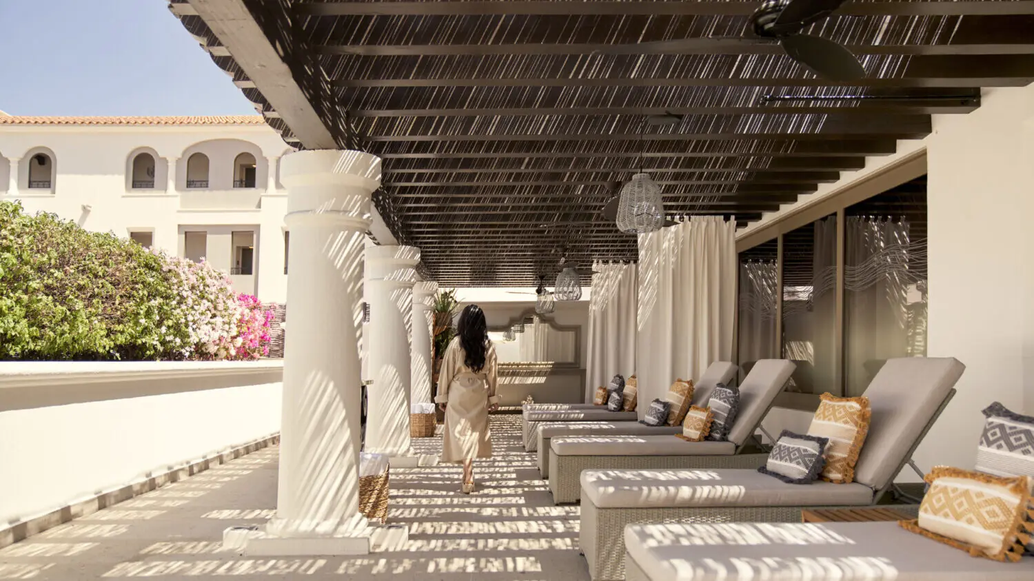 Guest walking through a shaded resort walkway with lounge chairs, white columns, and patterned sunlight.