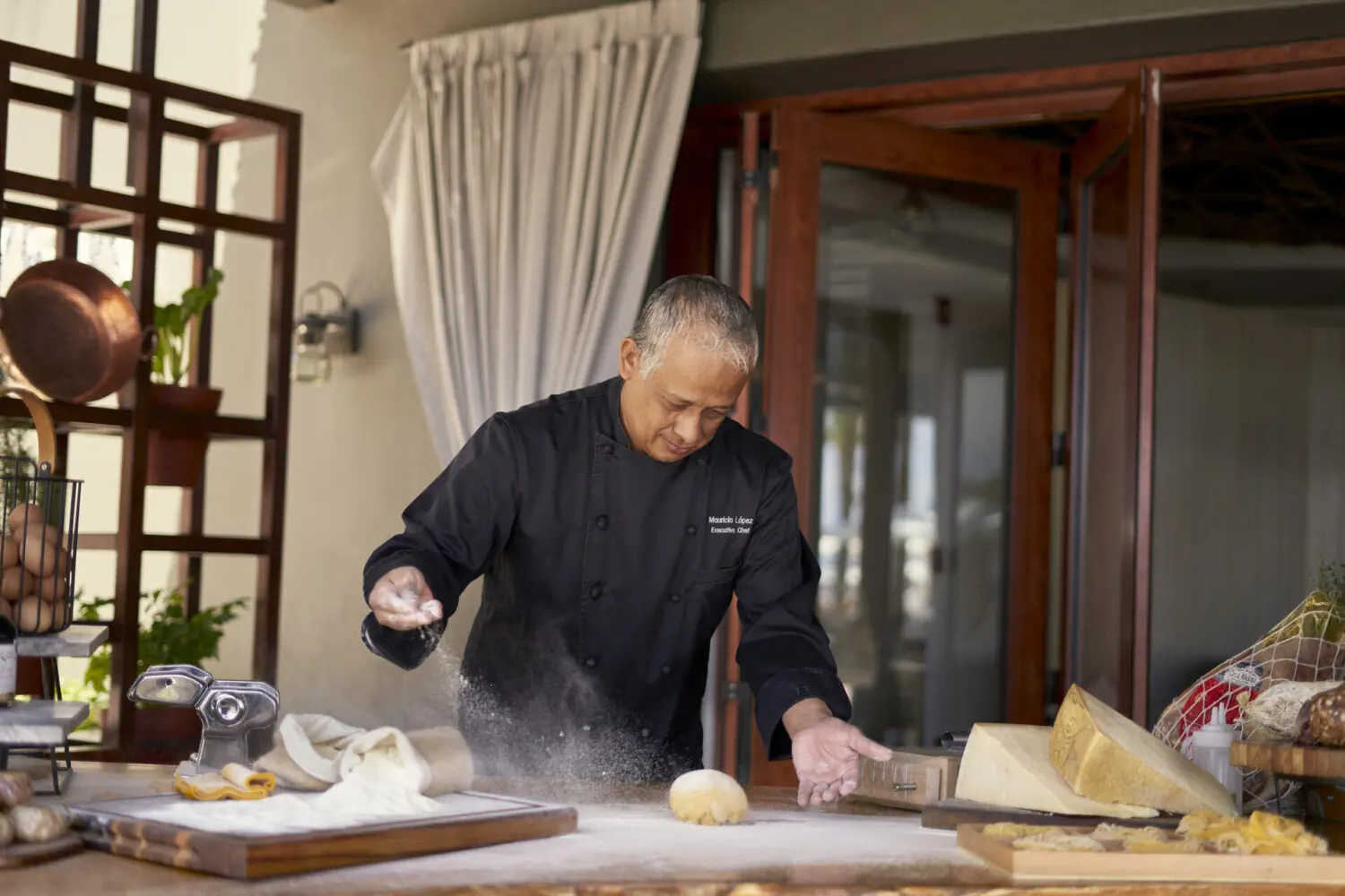 A chef dusts flour over fresh dough on a wooden table, surrounded by pasta tools and ingredients in a kitchen.