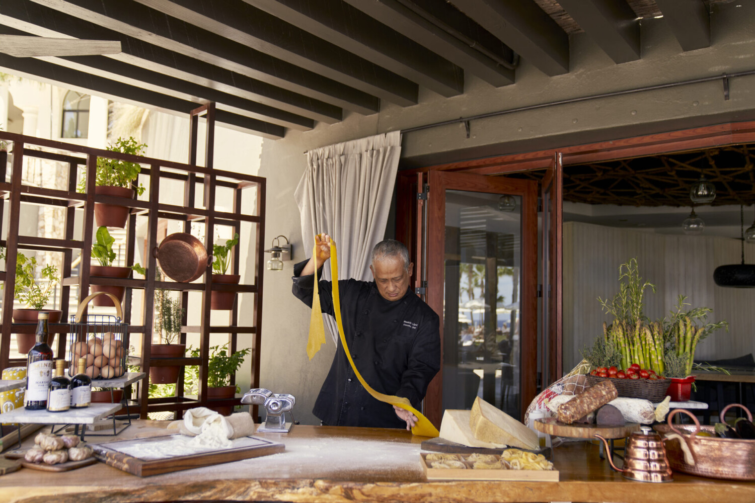 Chef preparing fresh pasta by hand at an outdoor kitchen with ingredients and cookware on display.
