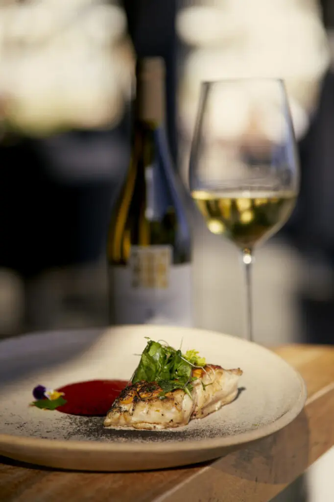 Plated grilled fish with sauce and greens on a table, paired with a glass and bottle of white wine.