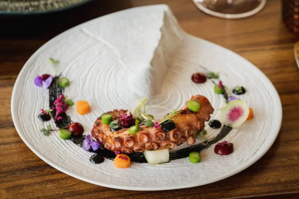 Plated gourmet octopus with colorful vegetables, edible flowers, and sauce on a textured white dish.