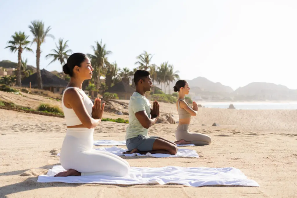 Three people practicing yoga on mats on a sandy beach with palm trees and ocean in the background.