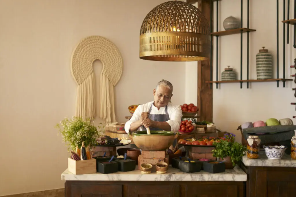 Chef in white shirt grinds ingredients in large bowl, surrounded by fresh produce and spices.