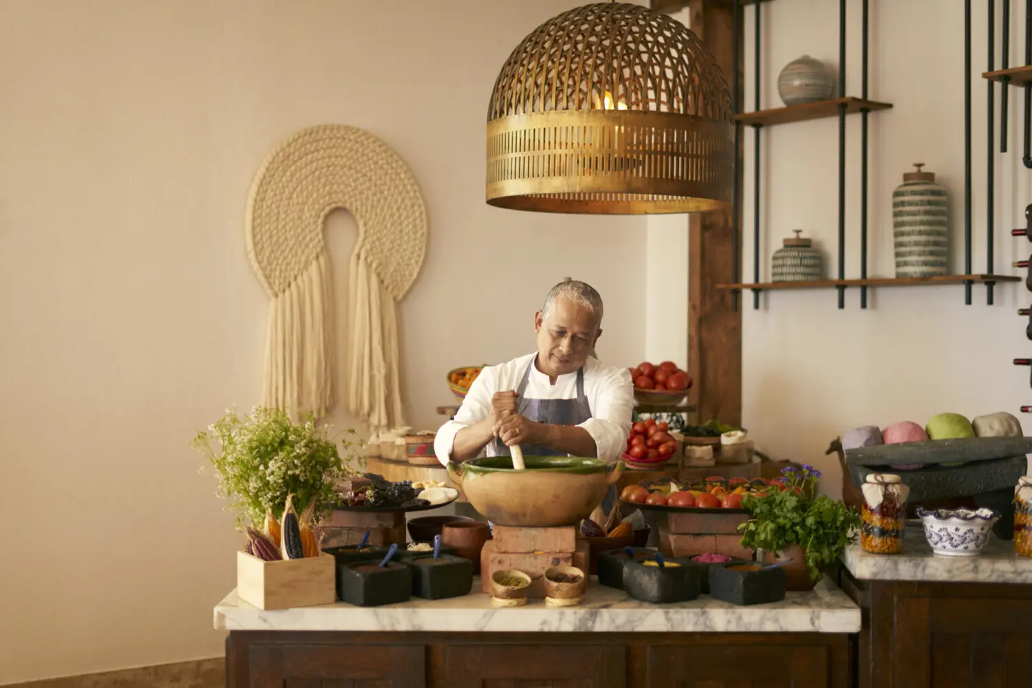 Un chef con camisa blanca muele ingredientes en un tazón grande, rodeado de productos frescos y especias.