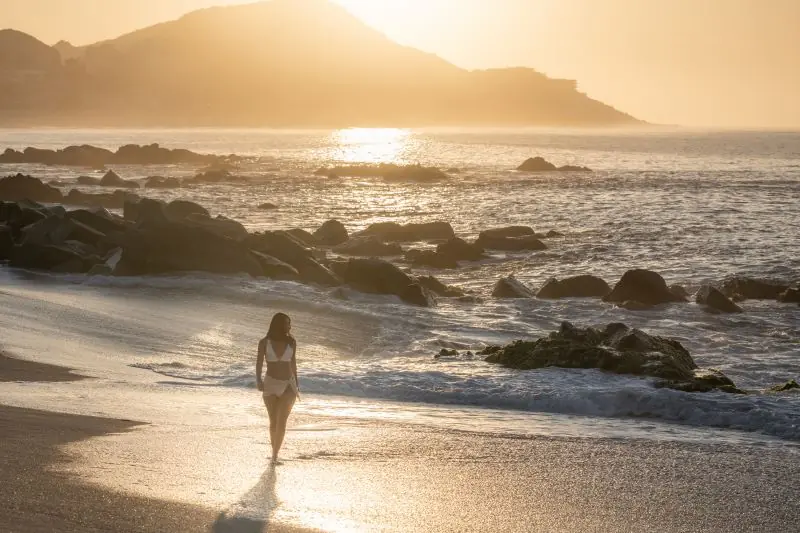 Person walking along a rocky shoreline at golden sunset with waves, mountains, and shimmering ocean light.