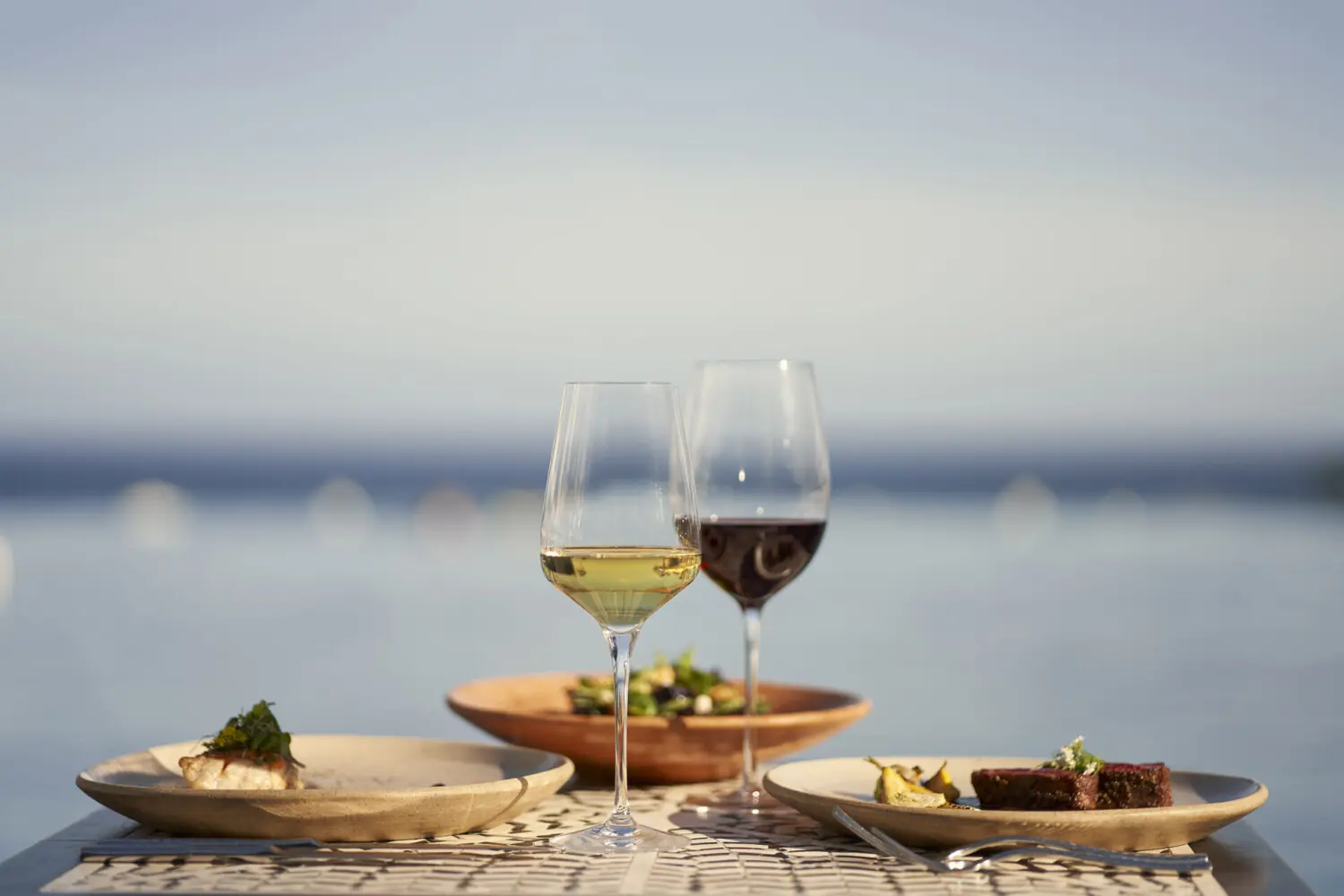Wine glasses and plated gourmet dishes set on an outdoor table overlooking calm water at sunset.