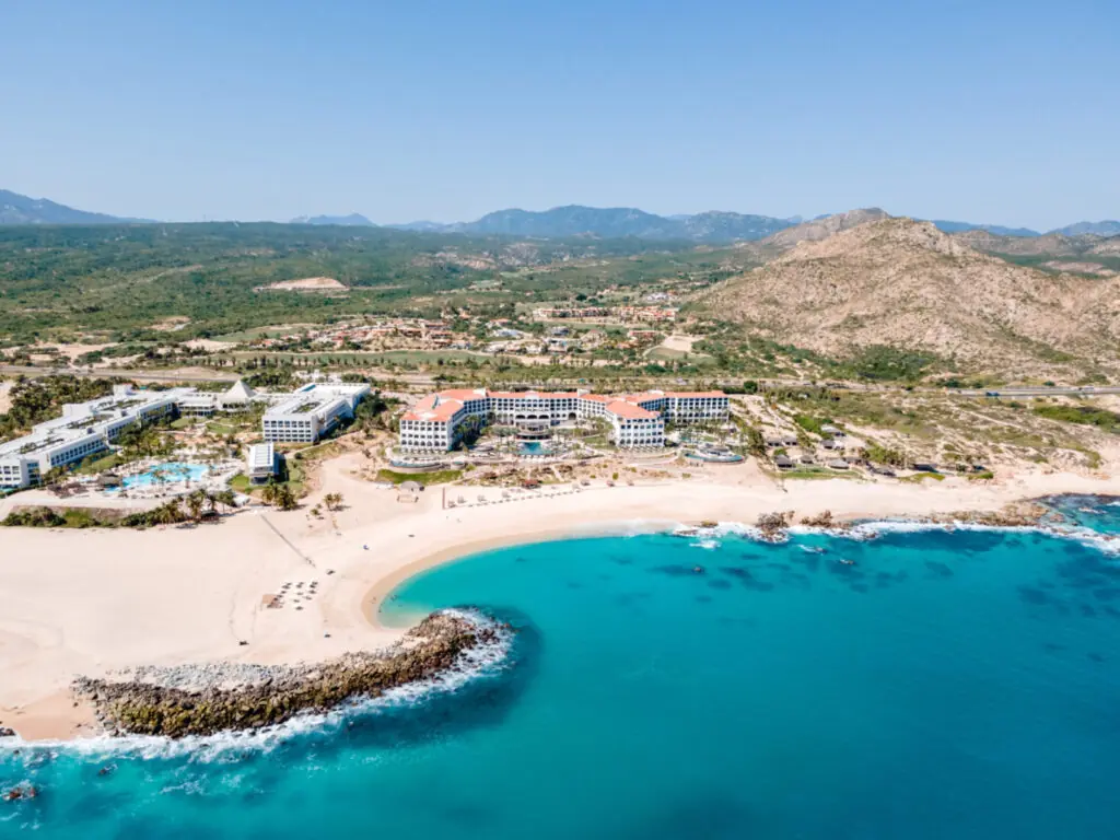 Aerial view of a beachfront resort with turquoise water, sandy shore, and mountains in the background.