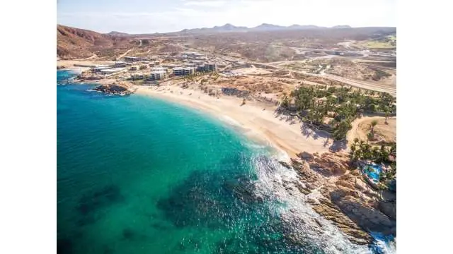 Aerial view of a turquoise beach cove with rocky cliffs, sandy shore, and coastal resort buildings nearby.