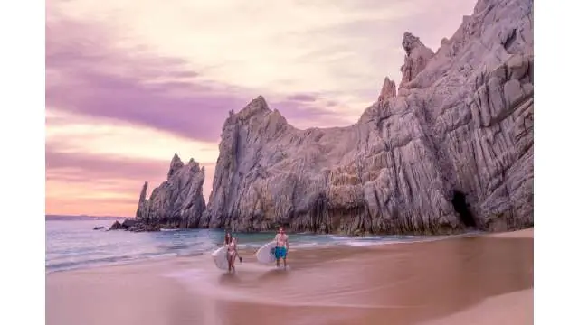 Surfers walking along a sandy beach with surfboards near dramatic rock formations at sunset.