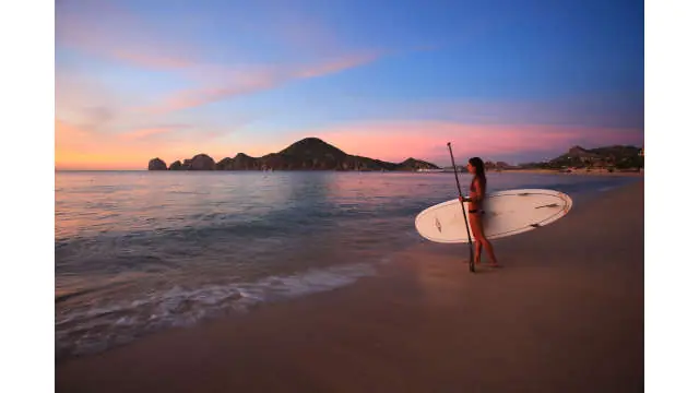 Person holding a paddleboard on a sandy beach at sunset, with calm ocean water and distant rocky hills.
