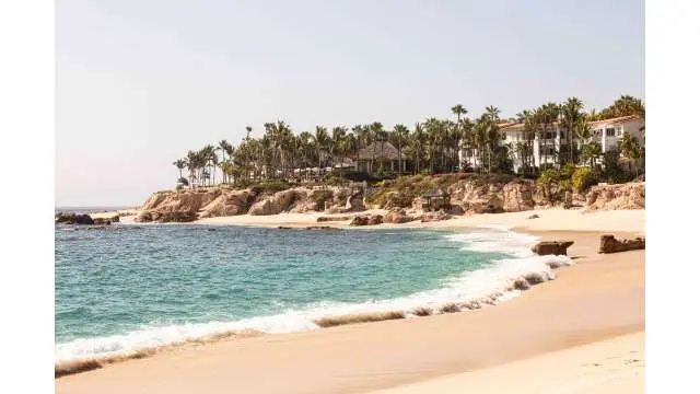 Sunny beach with turquoise waves, sandy shore, palm trees, and resort buildings along a rocky coastline.
