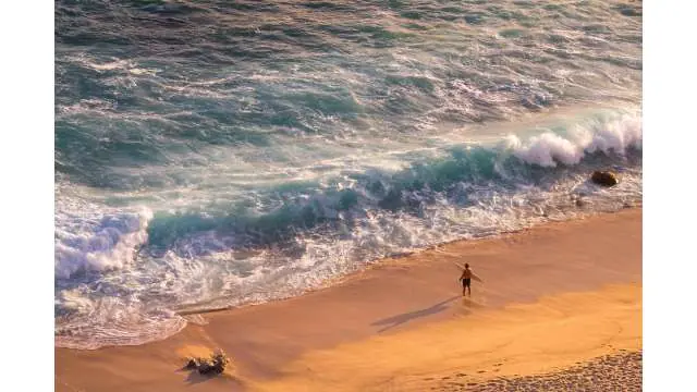Person walking alone along a sandy beach as waves roll in from the ocean, viewed from above.