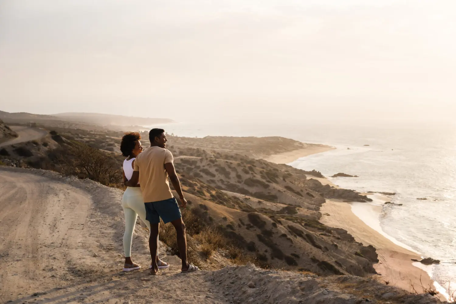 Couple standing on coastal cliff trail at sunset, admiring scenic ocean waves and sandy beach.