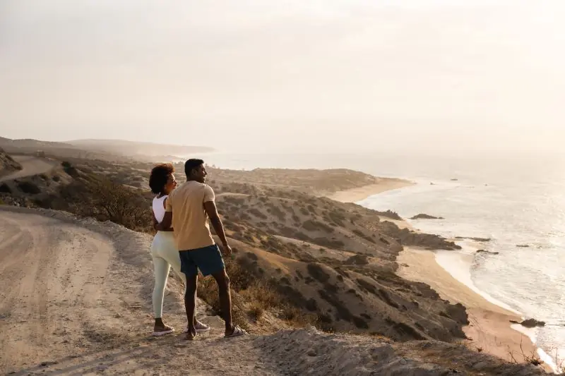 Couple standing on a coastal cliff overlooking the ocean and sandy shoreline during golden hour.