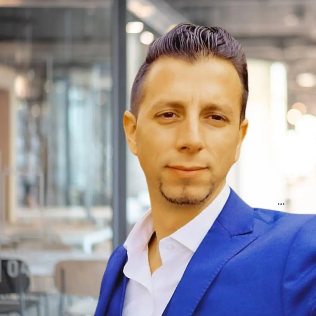 Man in a blue suit jacket and white shirt posing indoors in a modern office setting with glass walls.