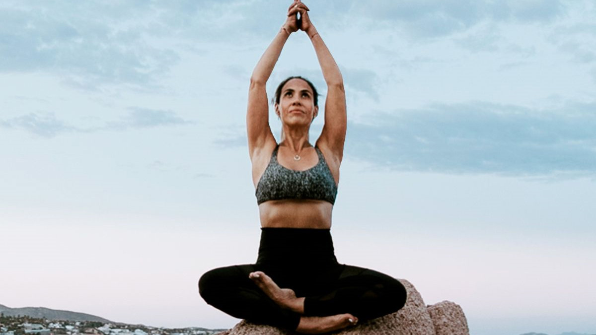 Woman practicing seated yoga pose on a rock outdoors, arms raised overhead against a calm sky backdrop.