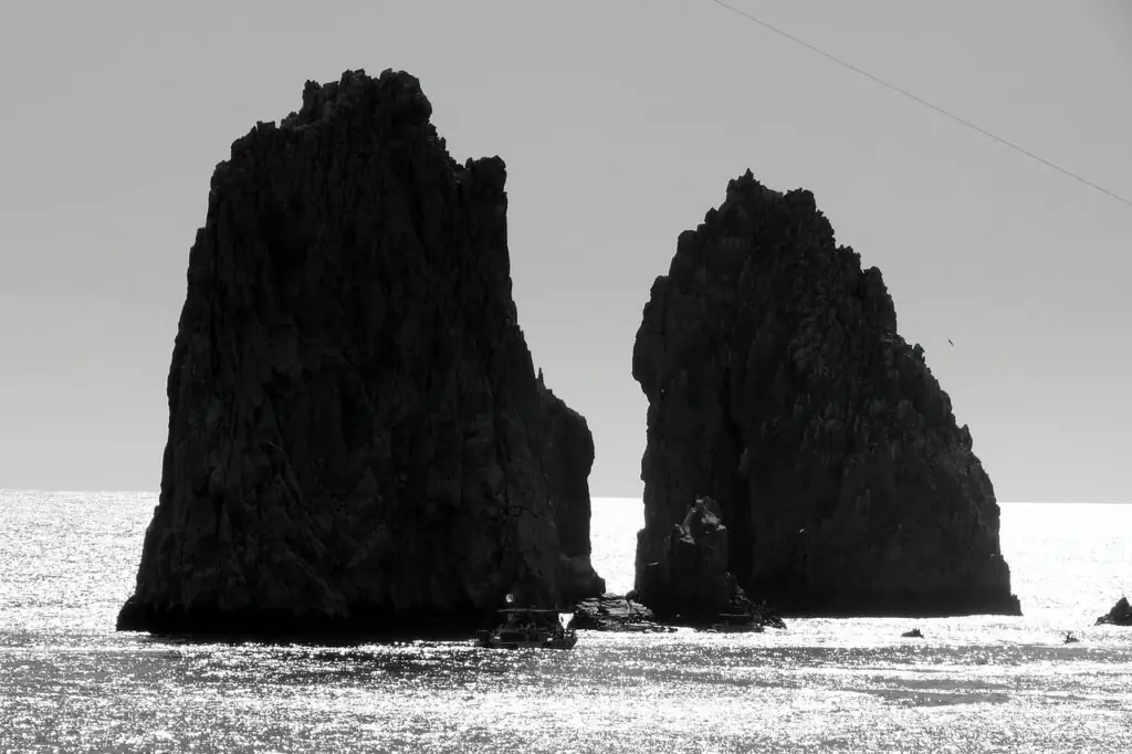 Two tall rock formations rising from the ocean, silhouetted against shimmering water and a bright sky.