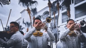 Mariachi musicians performing outdoors in traditional attire with papel picado banners and palm trees in the background.
