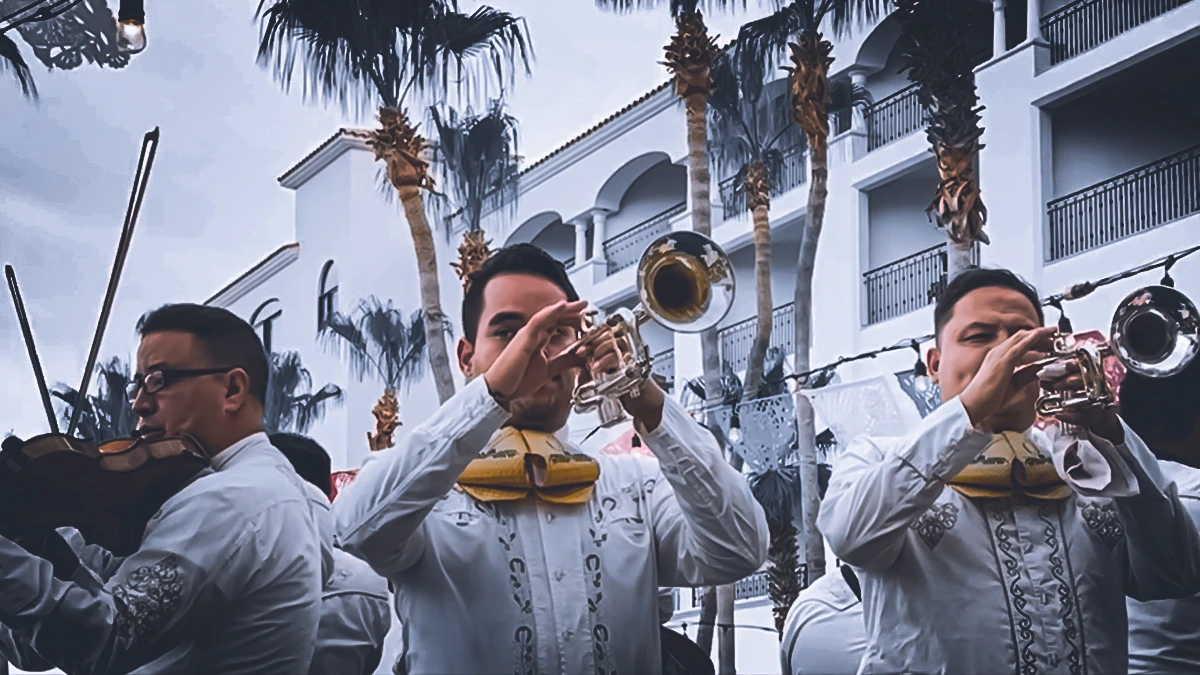 Mariachi musicians performing outdoors in traditional attire with papel picado banners and palm trees in the background.