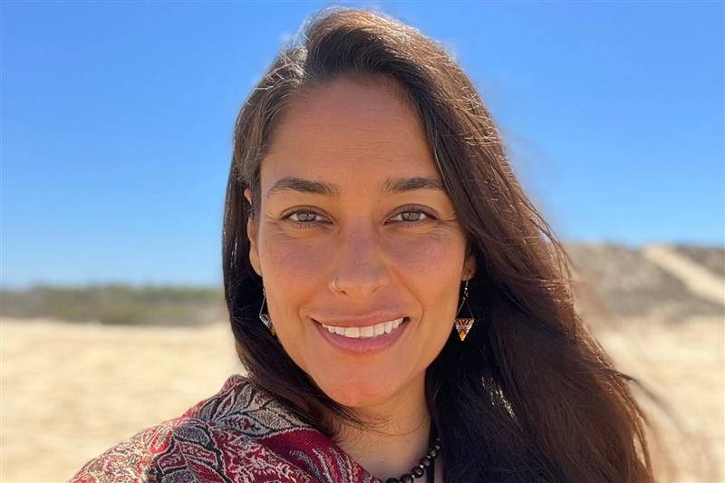 Mujer sonriente al aire libre bajo un cielo azul claro, vistiendo ropa estampada y aretes con un paisaje arenoso detrás de ella.