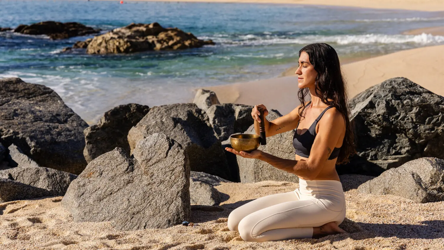 Person kneels on sandy beach near rocks, meditating and playing sound bowl by the ocean.